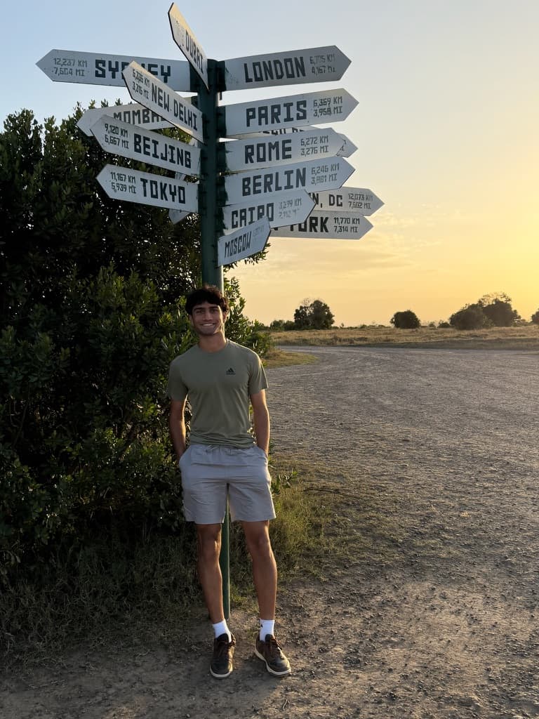 Amar standing by a world distances signpost at sunset